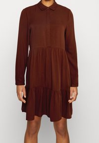 Robe marron à manches longues avec un col boutonné, présentant un ourlet à volants en étages et une texture lisse. Fabriquée en tissu léger.