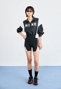 Veste coupe-vent noire et blanche de Nike avec fermeture éclair, associée à un short noir avec des accents gris. Le mannequin porte des lunettes de soleil et des chaussettes noires avec des chaussures.