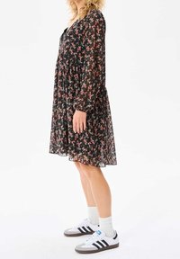 Nainen mustassa kukallisesa polvipitkässä mekossa, joka on yhdistetty valkoisiin Adidas-tennareihin ja valkoisiin nilkkasukkia, seisoo sivuttain valkoista taustaa vasten.