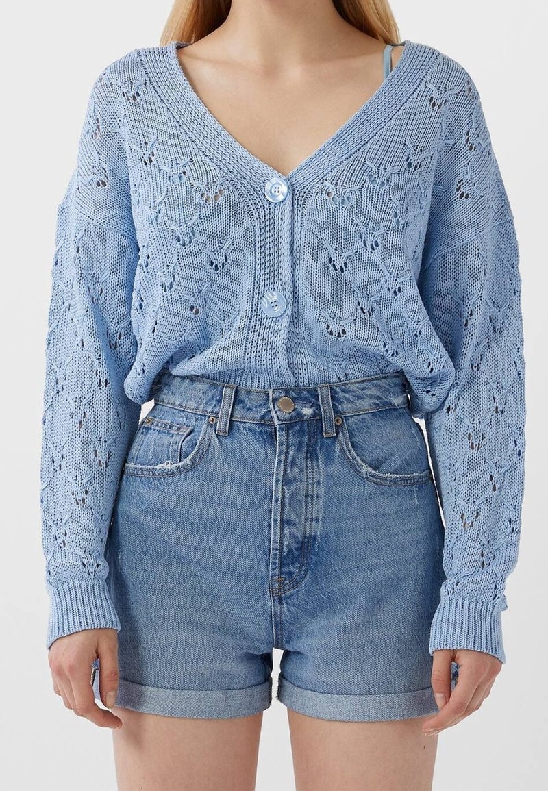 Světle modrý pletený cardigan s výstřihem do V, zdobený listovým vzorem a dvěma předními knoflíky, v kombinaci s džínovými kraťasy s vysokým pasem.
