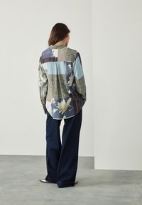 Chemise à manches longues avec un design color block en bleus, verts et bruns, présentant des motifs floraux et une coupe décontractée. Associée à un pantalon en denim à jambes larges.