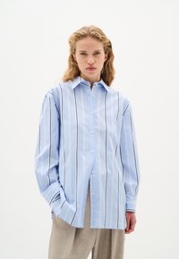 Camicia azzurro chiaro con bottoni, a righe verticali bianche e scure, maniche lunghe e colletto a punte; tessuto liscio e vestibilità comoda.