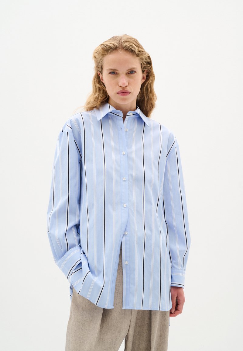 Camicia azzurro chiaro con bottoni, a righe verticali bianche e scure, maniche lunghe e colletto a punte; tessuto liscio e vestibilità comoda.