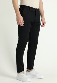 Pantalones negros ajustados de tela suave, con un corte slim, trabillas para cinturón, bolsillos laterales y cierre de botón, combinados con zapatillas blancas.