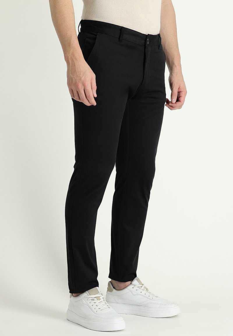 Pantalones negros ajustados de tela suave, con un corte slim, trabillas para cinturón, bolsillos laterales y cierre de botón, combinados con zapatillas blancas.