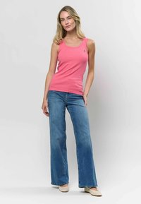 Rozā ribots bez piedurknēm tops ar plašām siksnām, kombinēts ar gaiši ziliem platiem džinsiem. Topam uz krūtīm ir neliels logotips. Modelis stāv pretī gaišai fonā.