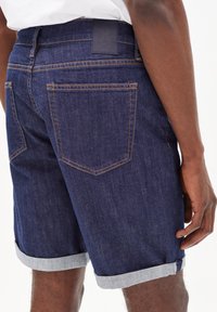 Shorts in denim blu scuro con orli risvoltati. Presentano una patch in pelle sul retro e cuciture arancioni a contrasto. Due tasche posteriori visibili.