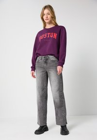 Lila Sweatshirt mit rotem Druck "BOSTON 1980", kombiniert mit hellgrauen geraden Jeans und schwarzen klobigen Stiefeletten.