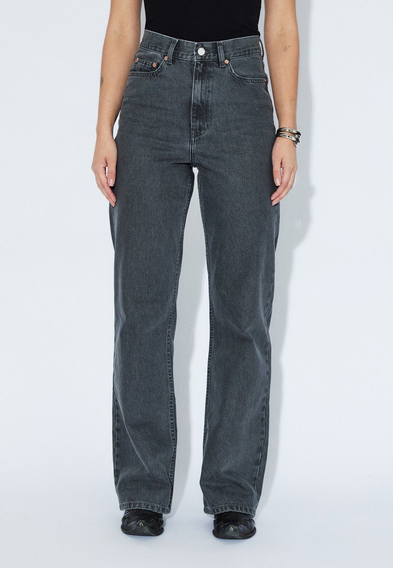 Frau trägt hoch taillierte, lockere dunkelgraue Jeans mit schwarzen Schuhen und einem schwarzen Oberteil, stehend vor einem schlichten hellen Hintergrund.