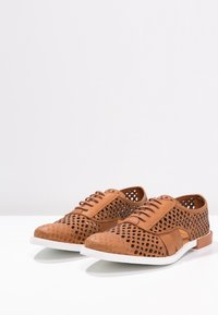 Scarpe in pelle perforata color cuoio con punta arrotondata, suola bianca e dettagli di design sottili, caratterizzate da trame e lacci a contrasto.