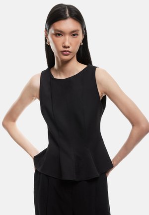 Top noir sans manches avec un buste ajusté, un ourlet en peplum et des coutures avant ; fabriqué dans un tissu lisse et texturé. Le mannequin pose avec les mains sur les hanches.