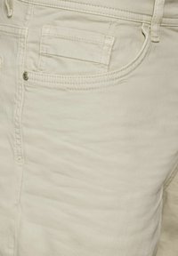 Krémové džíny s jemnou texturou, které mají přední kapsy, viditelný zip a kontrastní švy. Kovový knoflík jako ozdoba.