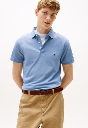 Fiatal férfi világoskék galléros ingben, fehér pólóval alatta, bézs nadrágban és barna övvel, kezeit a hát mögött tartva áll.