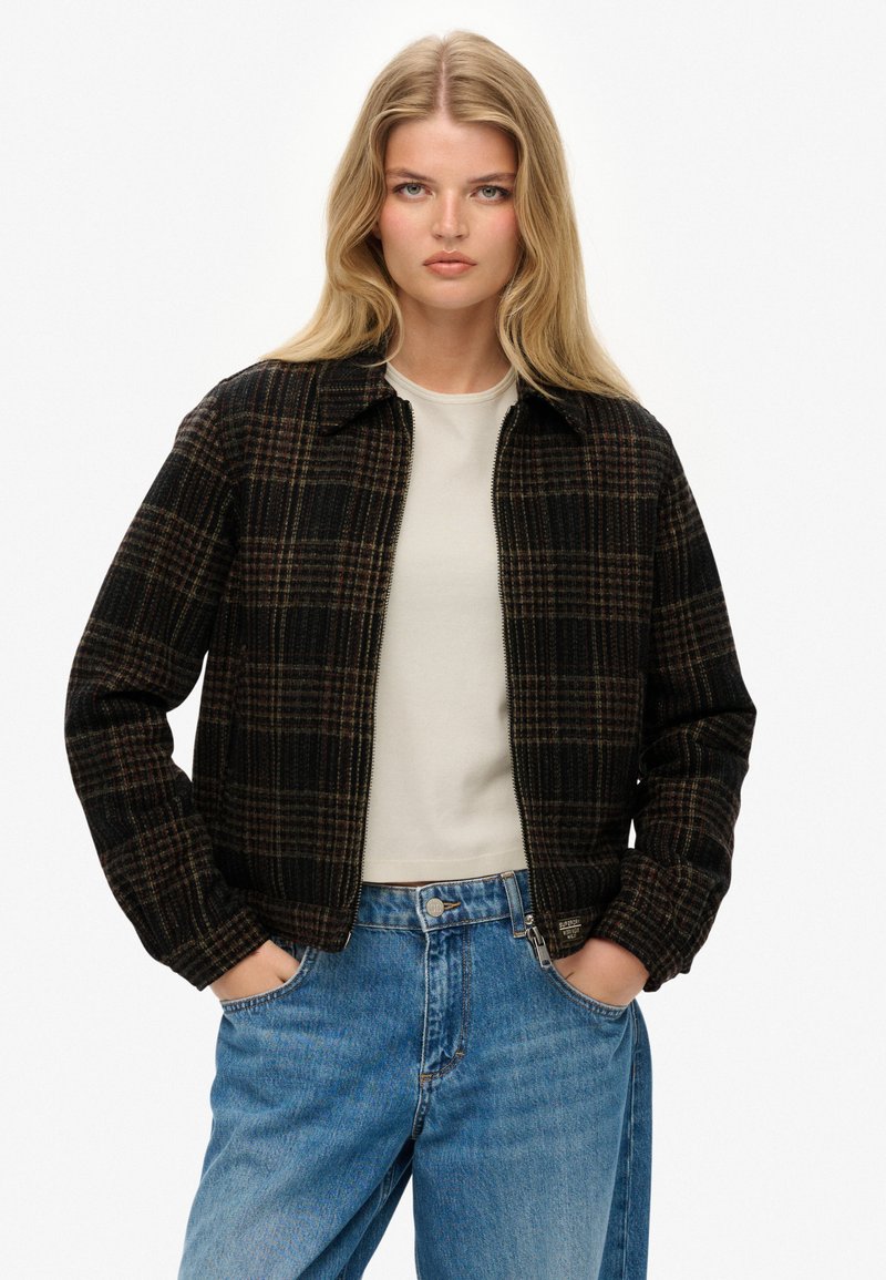 Geruite zwarte en bruine zip-up jack met een kraag en geribde mouwen, gecombineerd met lichtblauwe high-waisted jeans.