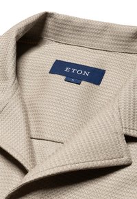 Tessuto beige texturizzato con un motivo intrecciato, caratterizzato da un'etichetta ETON blu cucita nel colletto e un colletto ripiegato.