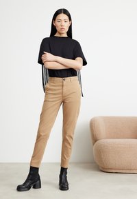 Černé tričko s krátkým rukávem a třásněmi, béžové tapered kalhoty a černé boty na podpatku. Měkké textury a neutrální barvy. Minimalistický design.
