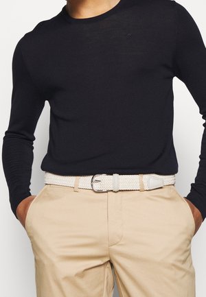 Homme portant une chemise noire à manches longues rentrée dans un pantalon beige avec une ceinture tissée blanche, les mains dans les poches.