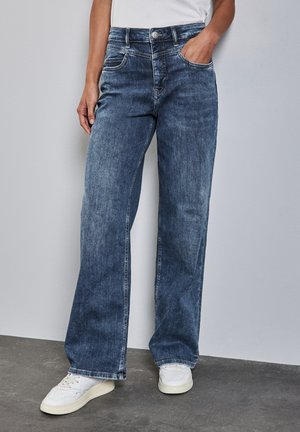 Wide-leg denim jeans in a medium blue wash with a faded texture, featuring five pockets and a button closure, paired with white sneakers.