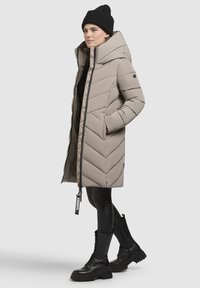 Beige puffer coat with a chevron pattern, large hood, zip closure, and two side pockets, paired with black leggings and chunky black boots.