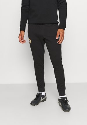 Mann trägt schwarze Jogginghose mit einem Sportlogo am Oberschenkel, kombiniert mit schwarzen Nike-Stollenschuhen und weißen Socken, steht auf einem grauen Boden.