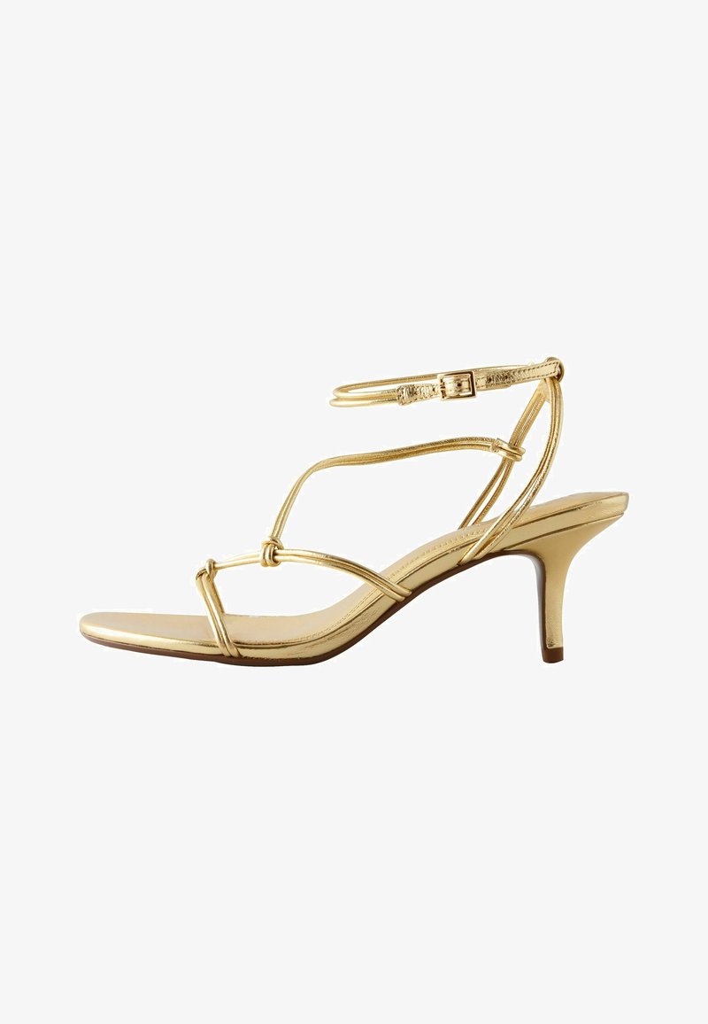 Sandale à talons hauts dorée avec des lanières fines, une boucle à la cheville et un talon moyen mince sur fond blanc.