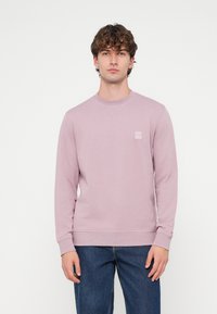 Svetlofialová mikina s dlhými rukávmi, okrúhlym výstrihom, jemnými detailmi švov a malou textilnou nášivkou na hrudníku. Nosí sa s modrými džínsami.