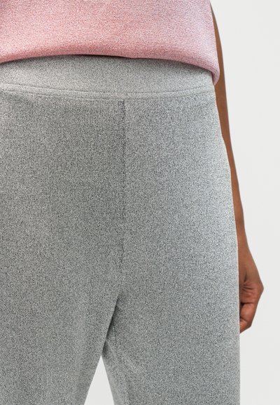 Gros plan sur une personne portant un pantalon en tricot gris et un haut rose, montrant la taille et la main droite sur un fond clair.