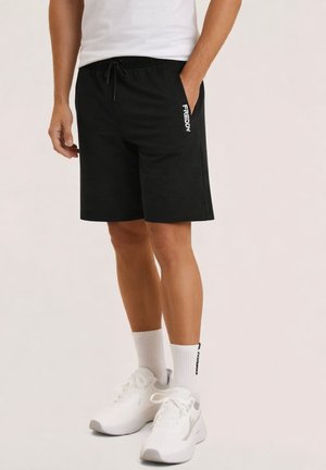 Persona che indossa pantaloncini neri del marchio Freddy, calzini bianchi con scritte nere e scarpe da ginnastica bianche, in piedi contro uno sfondo neutro.