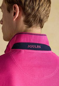 Roosa polo särk tõstetud kraega, millel on tumesinine aktsent ja roosa broderi "JOULES". Teksureeritud kangas ühtlase värviga.