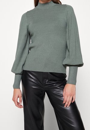 Femme portant un pull à col montant vert sauge avec des manches ballon et des poignets ajustés, associé à un pantalon noir en simili cuir.