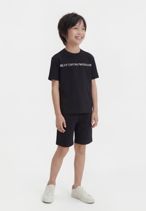 Mosolygó fiú áll, fekete EA7 Emporio Armani pólót, fekete rövidnadrágot és fehér tornacipőt visel, fehér háttér előtt.