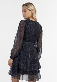 Lyhyt, tumma, monikerroksinen mekko, jossa on läpinäkyvät pitkät hihat, teksturoitu kangas, jossa on kukkakuvio ja kiristettävä vyötärö, jossa on nauha.