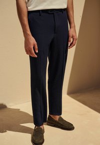 Pantalons textur�és bleu marine avec une coupe ajustée, dotés de poches latérales et d'une fermeture à bouton, associés à des mocassins vert foncé.