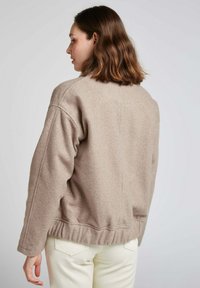 Veste longue beige texturée à manches longues avec une coupe décontractée, un col rond et un ourlet froncé, associée à un pantalon blanc.