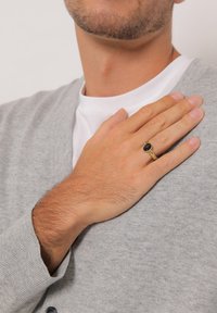 Guldring med en oval svart ädelsten, som bärs på en hand som vilar på en grå cardigan; huden visar lätt hår och en naturlig textur.
