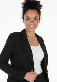 Czarna marynarka o dopasowanym kroju, z wyciętymi klapami i pojedynczym zapięciem na guzik, noszona na białej koszuli. Gładka tkanina, subtelny połysk.