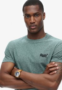 Groen gemêleerd T-shirt met ronde hals en korte mouwen, voorzien van een klein zwart logo op de borst. Gecombineerd met een gouden horloge en een tan kleurige leren band.