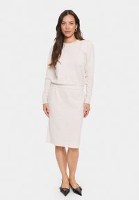 Hellbeige, langärmliges Kleid mit lockerer Passform, elastischem Bund, gerippten Bündchen und knielangem Saum; kombiniert mit schwarzen spitzen Schuhen.