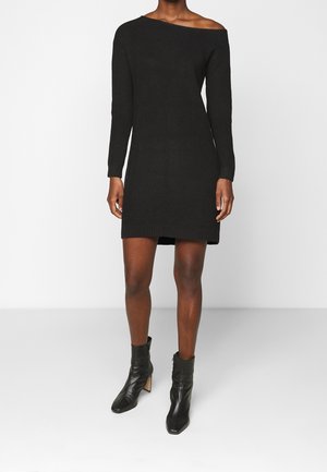 Woman wearing a black off-shoulder long-sleeve knit dress paired with black heeled ankle boots, standing against a plain white background.
