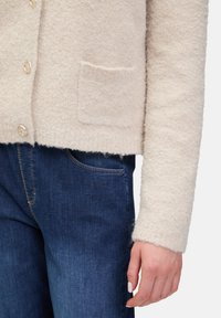Beige, texturerad cardigan med en framficka och guldknapplås, tillsammans med mörkblå jeans med sömnadsdetaljer.