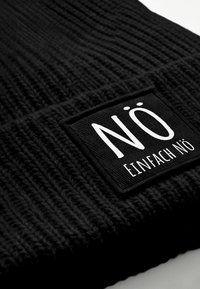 Black knit beanie with a textured pattern. Features a rectangular black patch with white text "NÖ" and "EINFACH NÖ." Soft material.