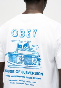 Πίσω μέρος λευκού πουκαμίσου με μπλε απεικόνιση κτιρίου, κείμενο «OBEY», «House of Subversion» και ονόματα πόλεων από κάτω.