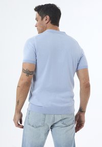 Polo de manga corta azul claro, hecho de tejido de punto, con cuello y dobladillo de canalé. Llevado con jeans de mezclilla claros; tatuajes visibles en los brazos.