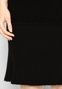Main à la peau claire posée sur une jupe noire en maille côtelée avec des motifs verticaux et diagonaux au niveau de la taille.