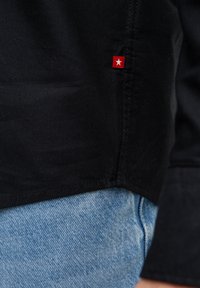 Crni rub košulje s crvenom oznakom na kojoj je bijela zvijezda, iznad svijetloplavih traperica i vidljiva manžeta na rukavu.