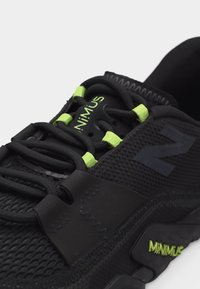 Μαύρο αθλητικό παπούτσι με πράσινες θηλιές κορδονιών, τη σήμανση "MINIMUS" στη γλώσσα και τη σόλα, και υφασμένο διχτυωτό και συνθετικό επάνω μέρος με υφή.
