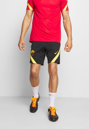 Mann in rotem Hemd und schwarzen Shorts mit gelben Akzenten, trägt weiße Socken und schwarze Fußballschuhe mit orangen Schnürsenkeln, steht auf grauem Boden.