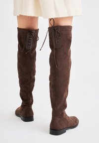 Botas de ante marrón hasta la rodilla con cordones en la parte posterior, llevadas con una falda de canalé crema, vistas desde atrás sobre un fondo claro.