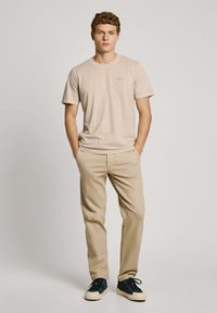 Camiseta de algodón beige con mangas cortas, combinada con pantalones beige. Los zapatos son oscuros con una suela blanca. Diseño simple y ajustado; sin patrones.