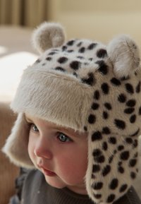 Plyšová čepice s leopardím vzorem a klapkami na uši, béžový podklad a chlupaté detaily. Měkká textura a zaoblené uši dodávají designu detaily.